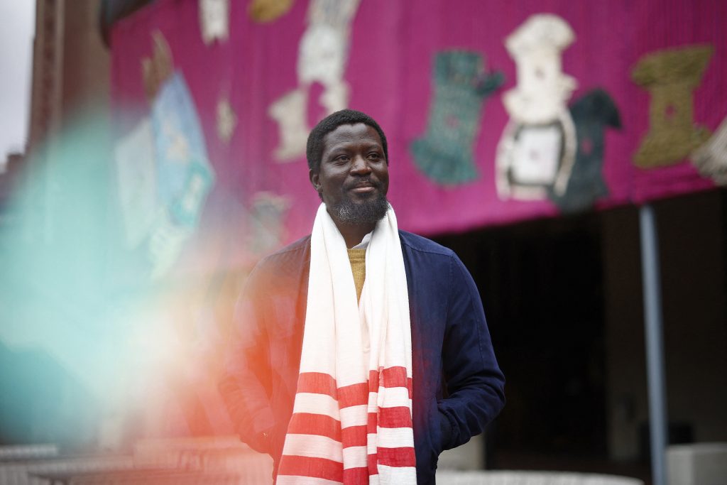 a man wearing a white scarf with red stripes stands in front of a large pink textile