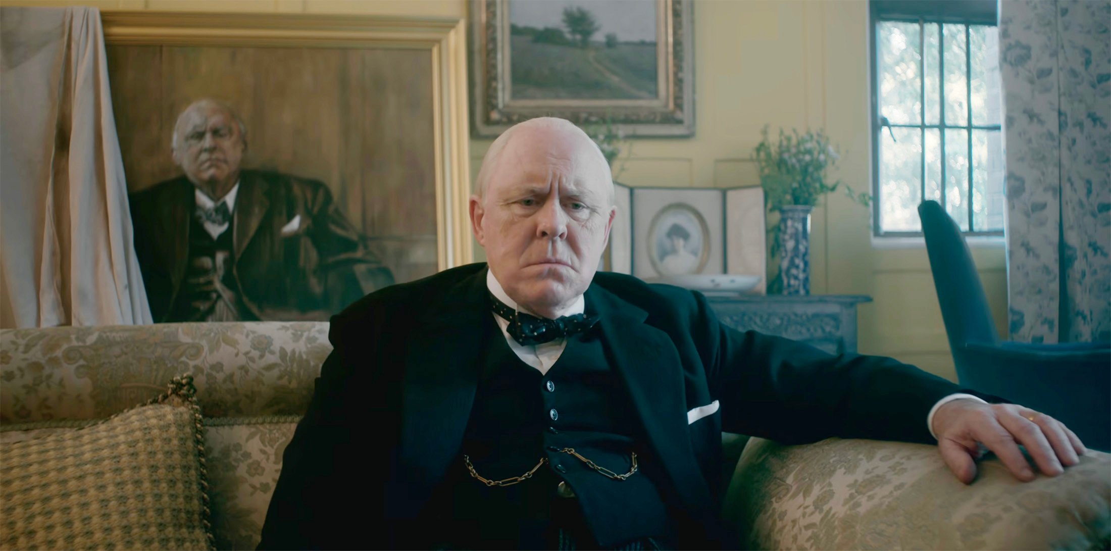 An elderly man sitting glumly on a sofa. Behind him is a portrait of him looking equally glum.