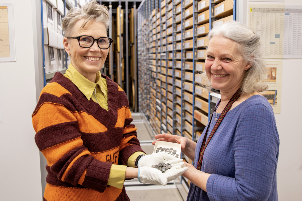 A photograph of two women in a museum archive room smiling and holding a single white gloved handful of very old silver coins