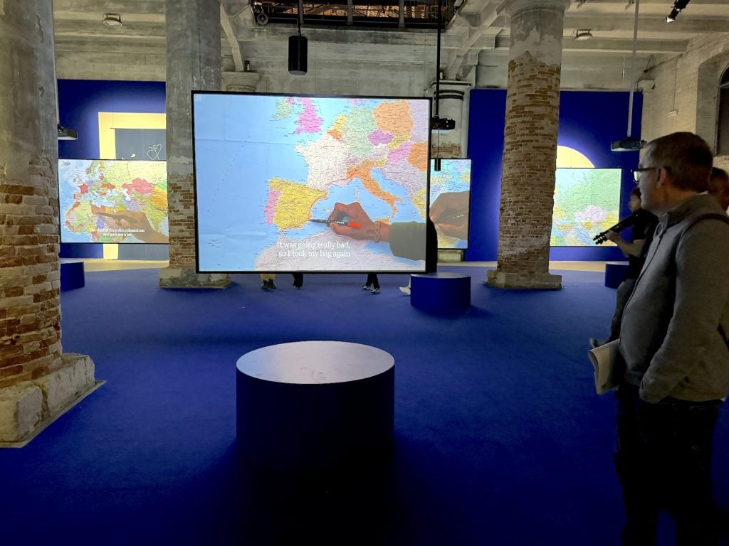 A man looks at an artwork consisting of multiple hanging screens showing hands writing on maps of North Africa and Europe