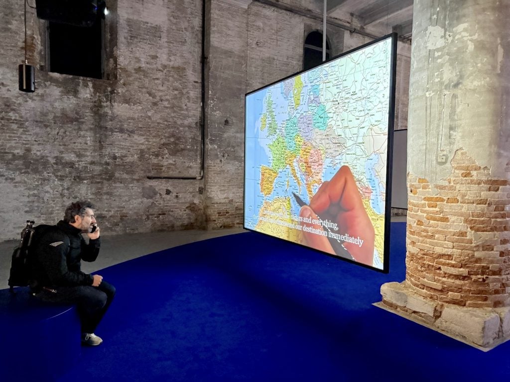 A man on a cell phone watches a large screen where a hand writes on a map of North Africa