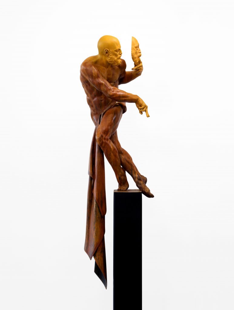 Sculpture of a nude man in motion with a strip of cloth around his waist and holding up a mask in front of his face.