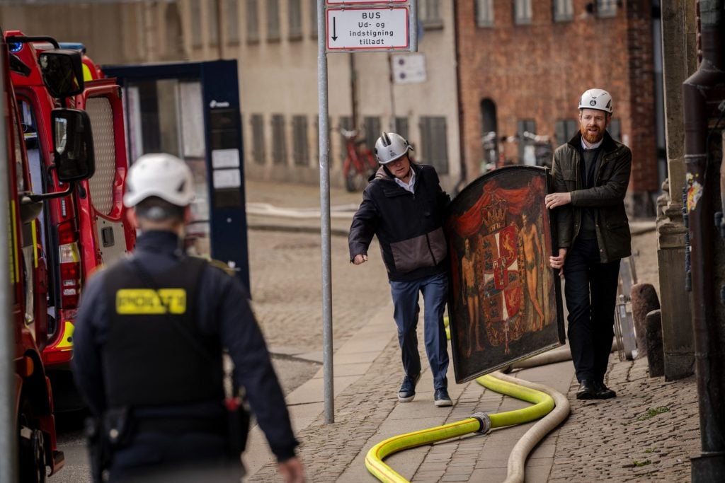 Two men transporting a painting on a street, while a fire truck idles on their right.