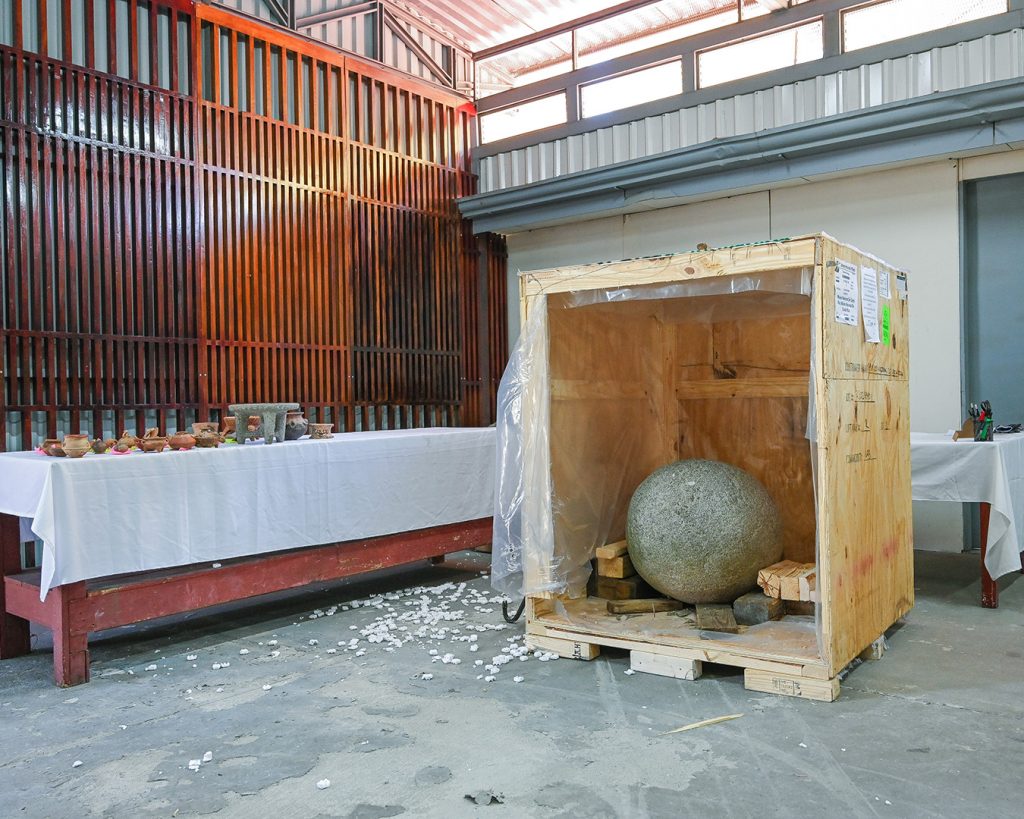 A photo of archaeological artifacts spread out across a table with a white cloth covering in a court yard, alongside an open wooden crate with a stone sphere in it