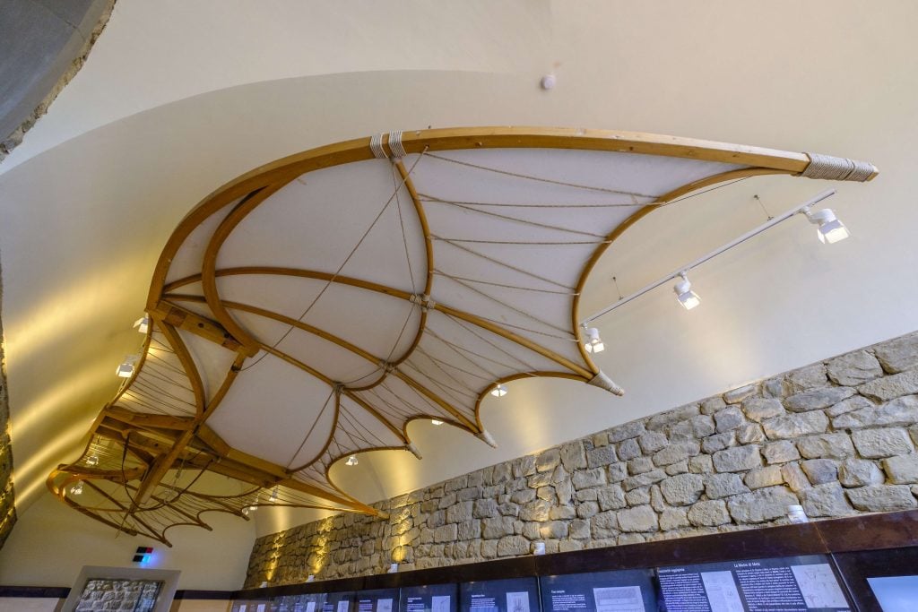 A photograph of Da Vinci's 15th century prototypical hang glider made from wood and displayed hanging on an angled white ceiling