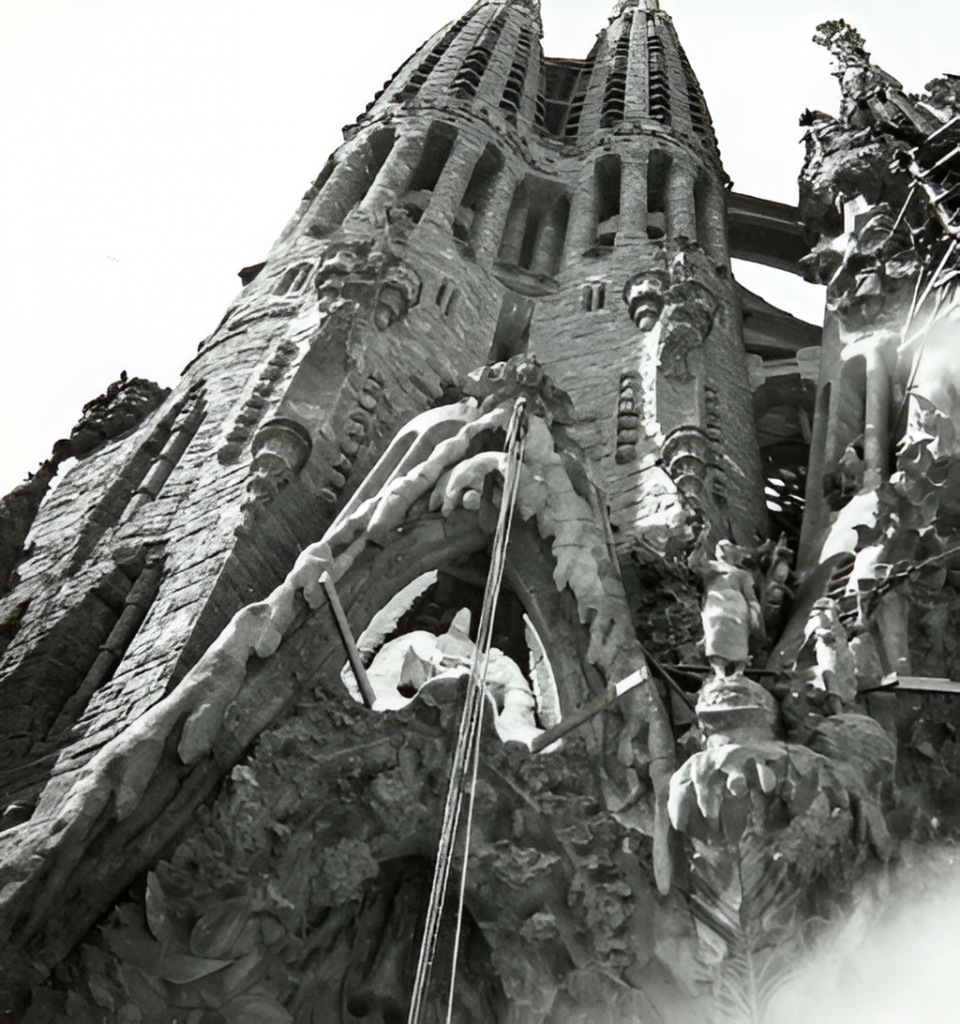 A black and white photograph of the Sagrada Família's Nativity façade, displaying its complex Gothic and Art Nouveau architectural details with sculpted figures and spires reaching upwards.