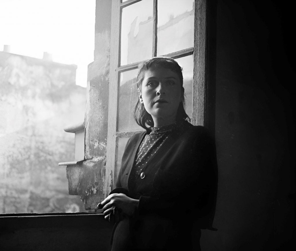 A black and white photograph of a woman standing by a window. Her expression is pensive and serious, directed outside the frame, suggesting contemplation or anticipation. The woman wears a dark, vintage-style dress with a dotted pattern, typical of mid-20th century fashion, adorned with a brooch. Light from the window illuminates her face and dress, creating a contrast with the darker interior. The urban landscape seen through the window's glass is blurred, giving an impressionistic feel to the background. The photo captures a quiet, introspective moment with a timeless quality.