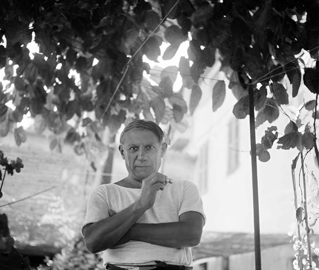 A black and white photograph of a middle-aged man standing under the foliage of a tree. The man has a contemplative expression, holding a cigarette between his fingers, bringing it close to his face. He wears a casual white T-shirt and appears relaxed yet thoughtful. The leaves create a dappled light effect, with patches of brightness highlighting parts of his figure and the surrounding environment. The background features a faint outline of a building, suggesting a tranquil domestic or garden setting. The composition captures a moment of quiet introspection in a natural setting.