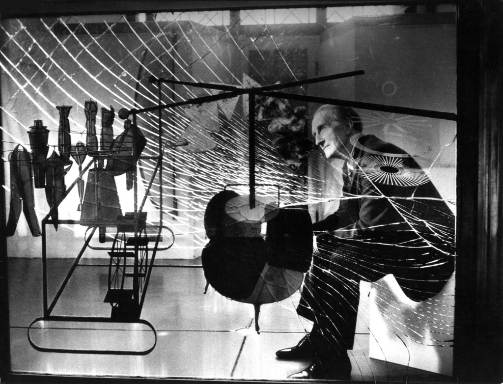 A black-and-white photo of the artist Marcel Duchamp sitting behind his abstract sculpture