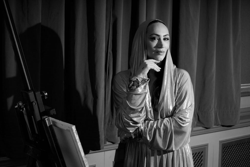 This is a black-and-white photograph of a woman dressed in a metallic-looking, shiny, silver hooded gown with a glamorous vibe. She is seen standing with her hand on her chin, possibly in a moment of thought or contemplation. She is positioned beside an easel, hinting at an artistic setting.