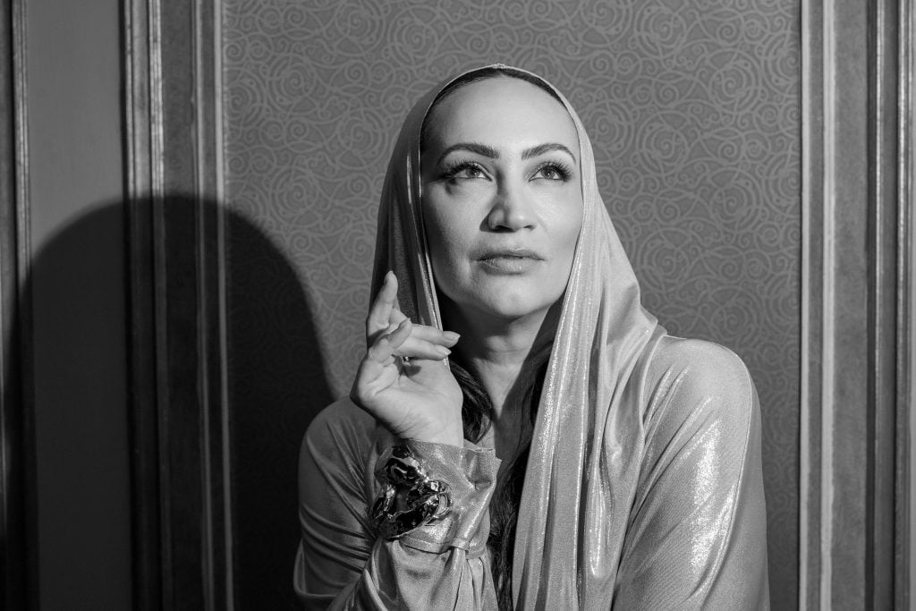 This is a black-and-white photograph of a woman dressed in a metallic-looking, shiny, silver hooded gown with a glamorous vibe. She is pictured with a thoughtful expression, her finger placed gently on her cheek, as she stands against a patterned wall. The pose and setting are reminiscent of classical portraiture, suggesting an air of elegance and introspection.