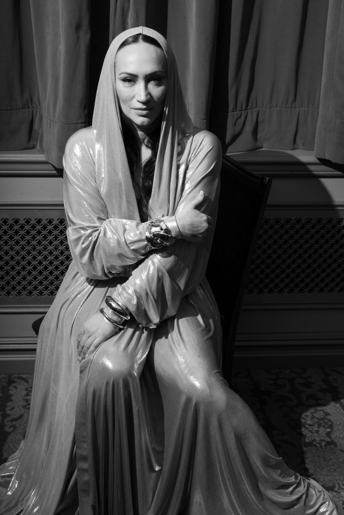 This is a black-and-white photograph of a woman dressed in a metallic-looking, shiny, silver hooded gown with a glamorous vibe. She is seen sitting down with crossed arms, exuding a sense of confidence and poise. Her posture and the direction of her gaze give the impression of a posed portrait.
