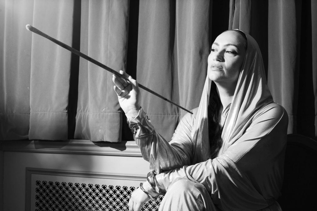 This is a black-and-white photograph of a woman dressed in a metallic-looking, shiny, silver hooded gown with a glamorous vibe. She is seen seated with a paintbrush in hand, looking at it intently. This suggests a connection to painting or artistry, and she appears to be either preparing to create art or in the midst of reflecting on her work.