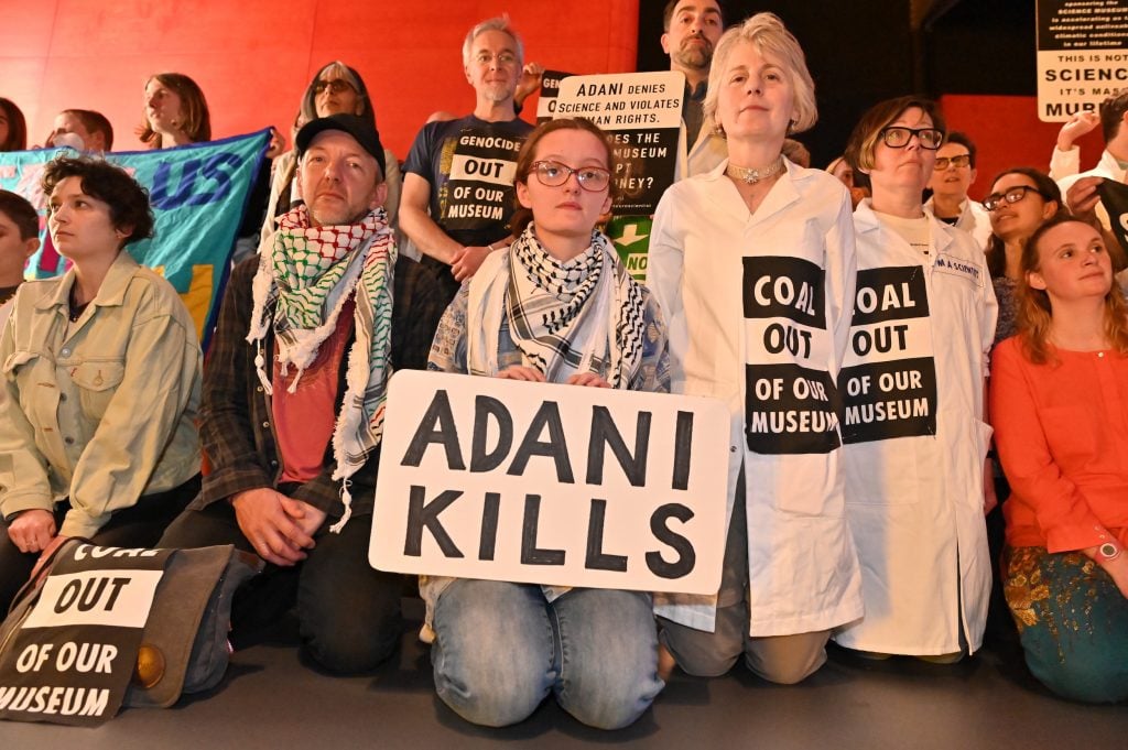 A group of people sitting and kneeling on the floor. One holds a protest sign that reads "Adani Kills."