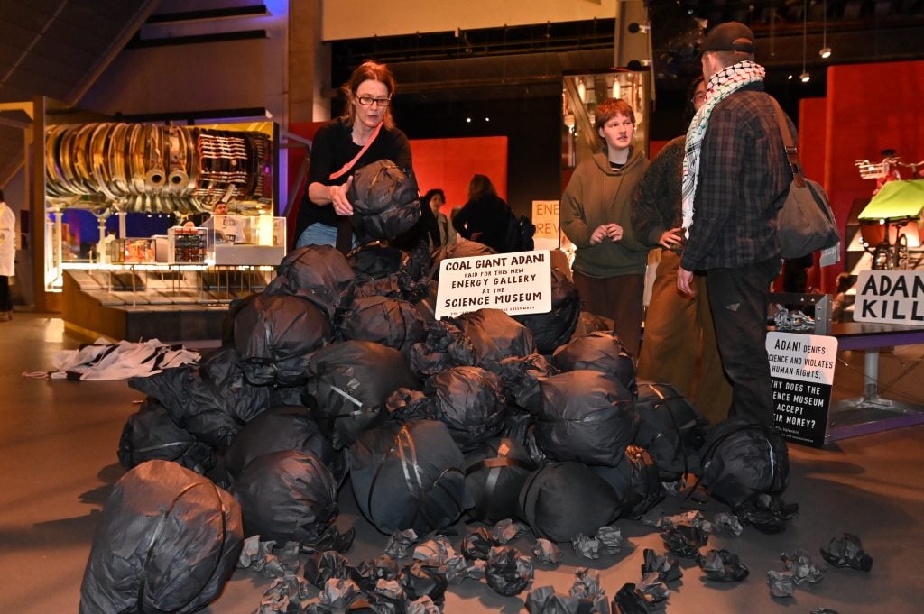 Black papier mache shaped like coal lumps sit in a heap on the floor. A woman is placing another lump on top of the pile.