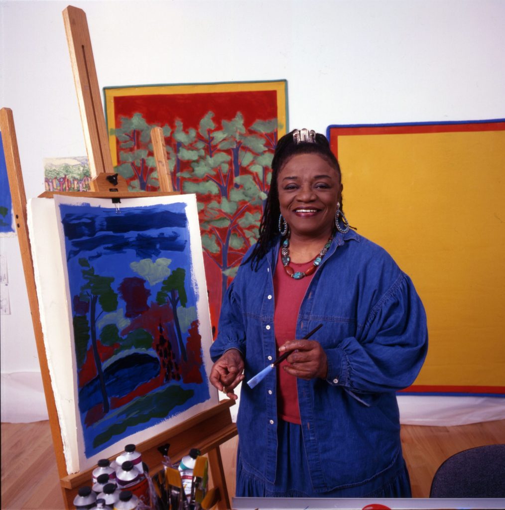 A woman, artist Faith Ringgold, standing in her studio beside one of her paintings, a paintbrush in hand.