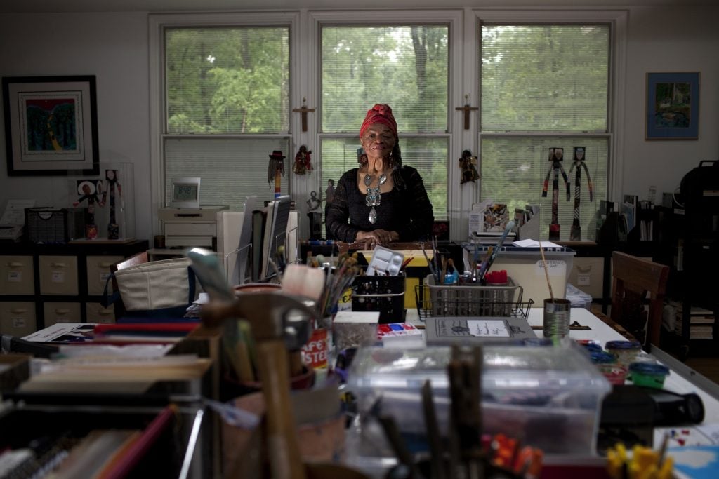 A woman, artist Faith Ringgold, standing at the end of a long table stacked with various art materials.