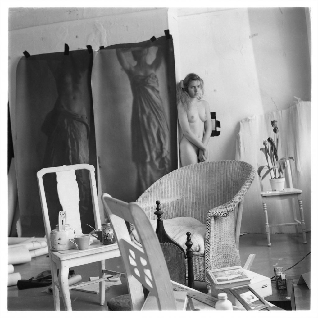 in a room cluttered with chairs, a woman stands in naked. She stairs out directly at the camera. Beside her taped to the walls are two monumentally scaled photographs of women posing as caryatids