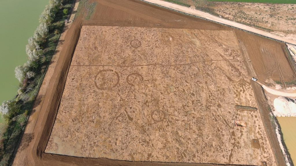 An aerial view of a dirt trodden excavated expanse lightly imprinted with five circular enclosures