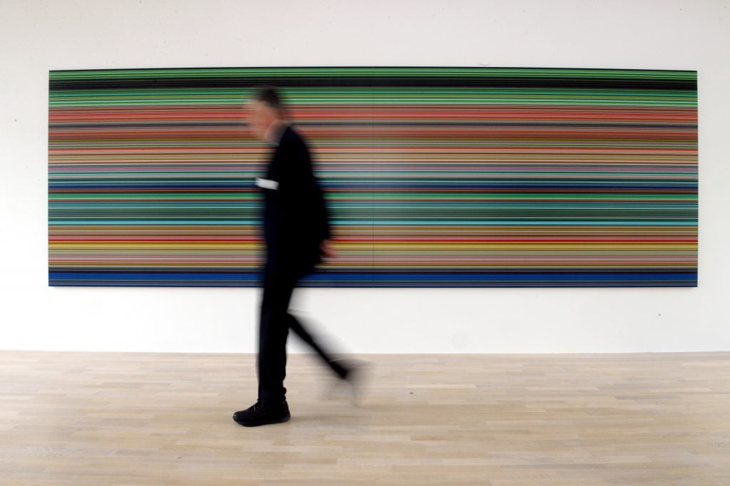 a blurry figure walks past a long horizontal painting with horizontal stripes running along it in different colours