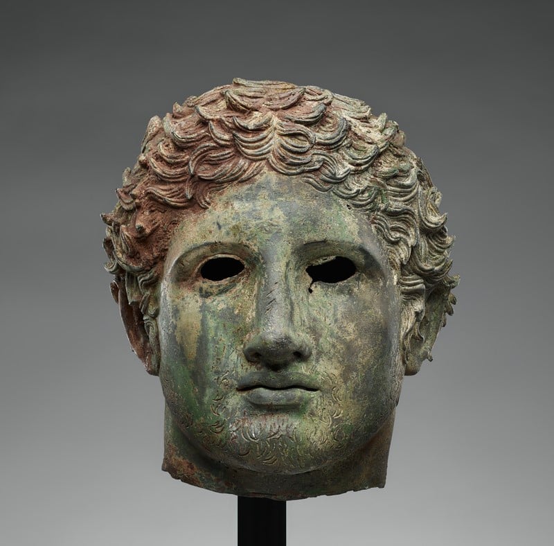 An ancient bronze head sculpture with a green patina, suggesting age and exposure to the elements. The sculpture, which appears to be of Greek or Roman origin based on the style of the hair and facial features, has intricate curls that are well-defined and cover the head. The eyes are hollowed out, possibly once inlaid with precious materials or stones to imitate a lifelike appearance. The face exhibits classical features: a straight nose, full lips, and a serene expression. The craftsmanship showcases the high level of artistry achieved in antiquity. The head is mounted on a simple, modern stand, which contrasts with the ancient artifact and emphasizes its historical significance.