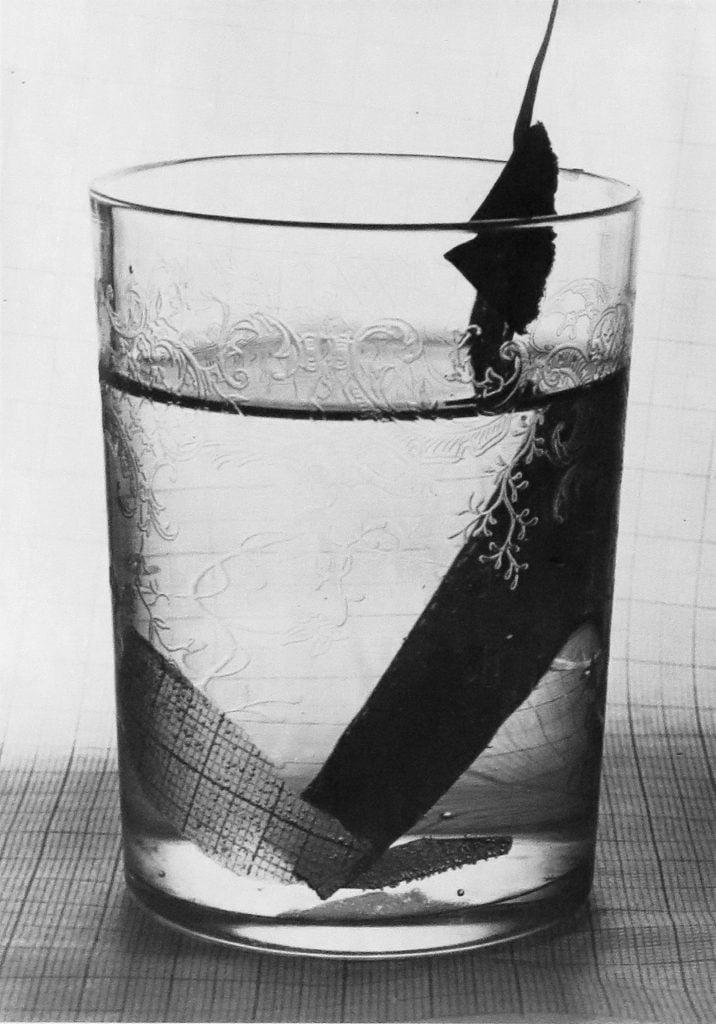 A black and white photo of a patterned clear glass with a strip of paper nearly submerged inside, to be shown at the Photography Show.