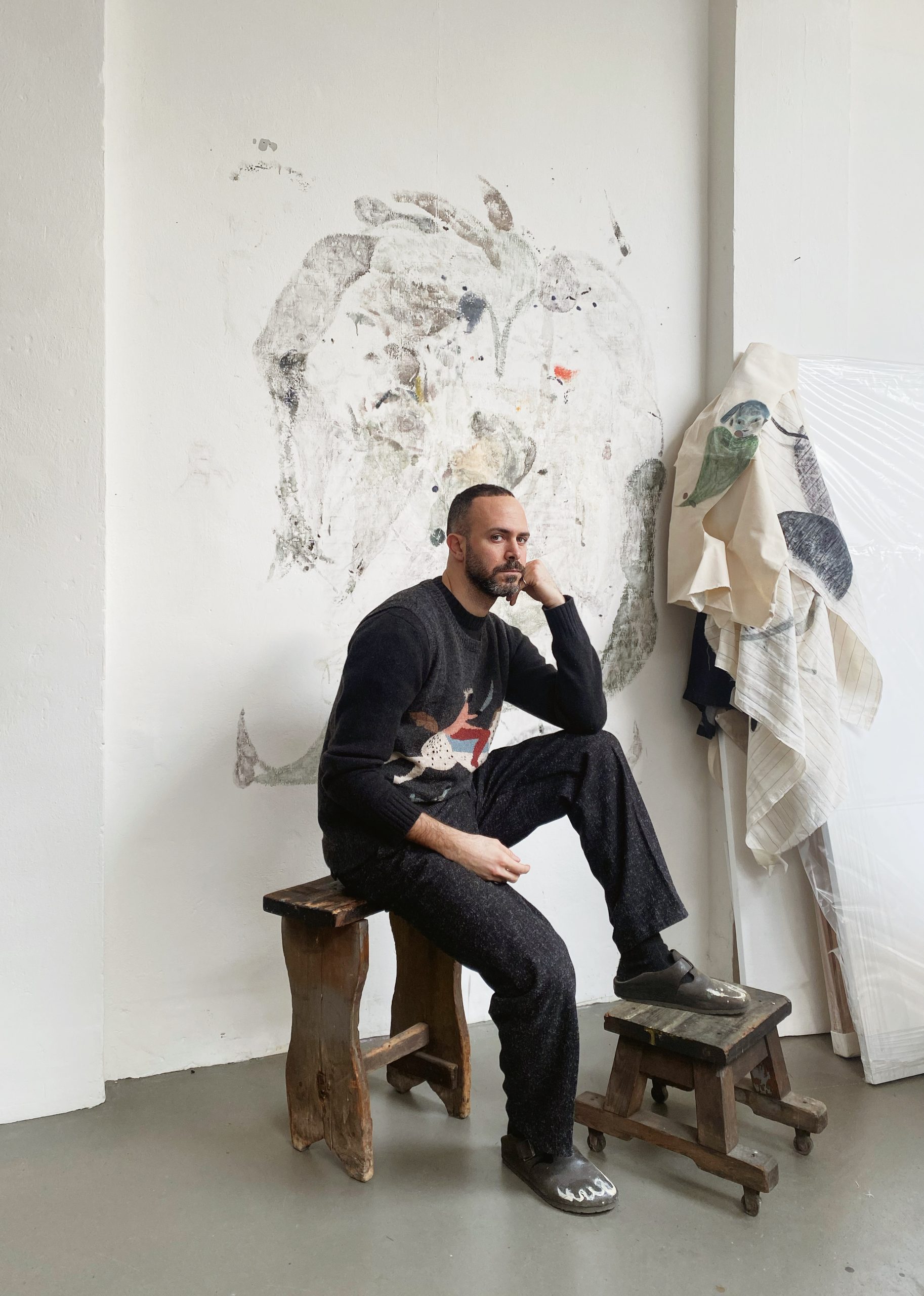 photograph of a white in a dark shirt and pants sitting on a bendh with his knee up. Beside him is a sculputre and behind him is a sketch-like painting