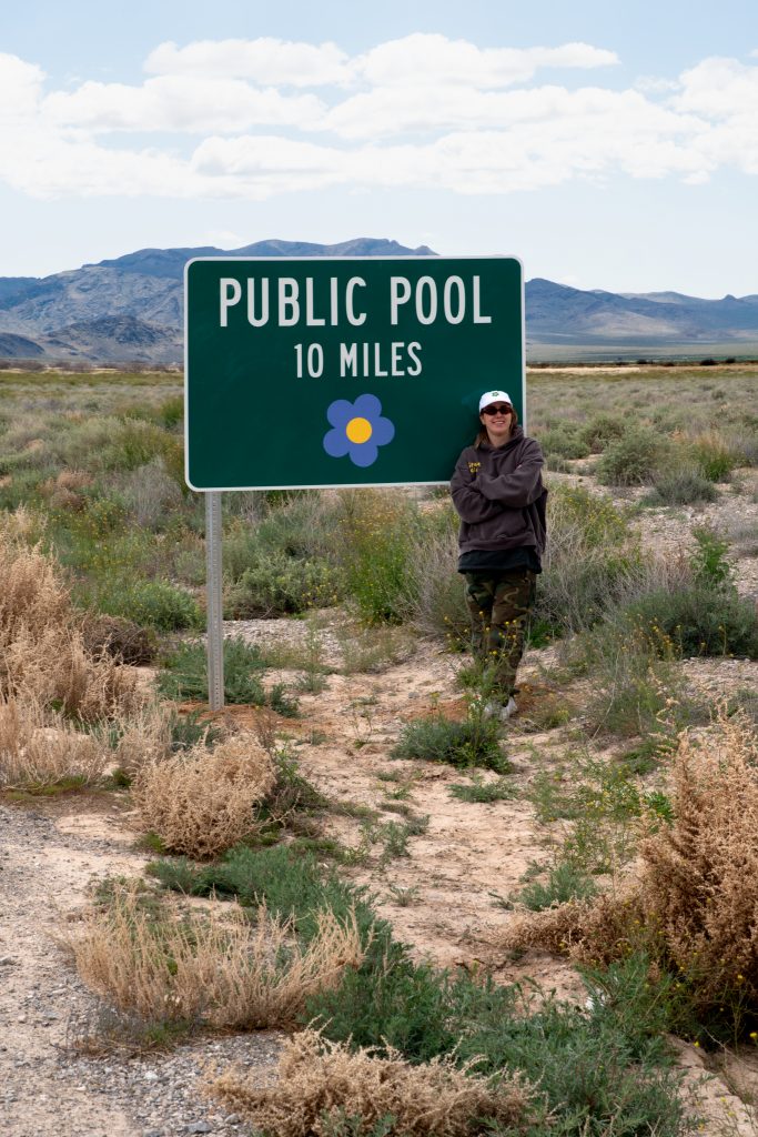 A photograph of a girl standing roadside before a mock highway sign amongst desert tumbleweeds