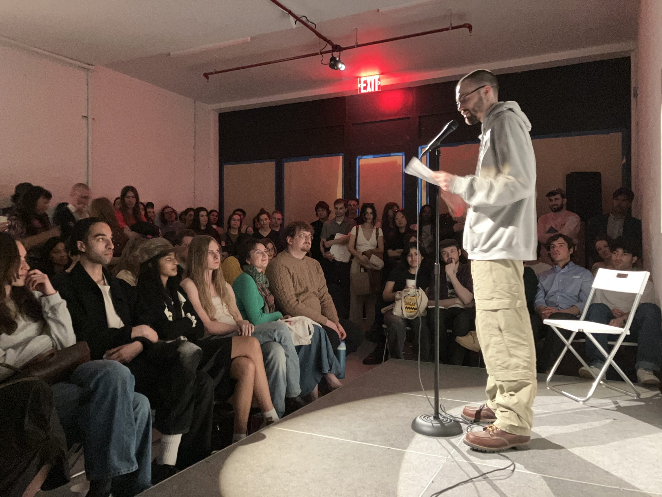 A photograph of a man standing on stage reading at a microphone for a crowd seated beneath red lighting