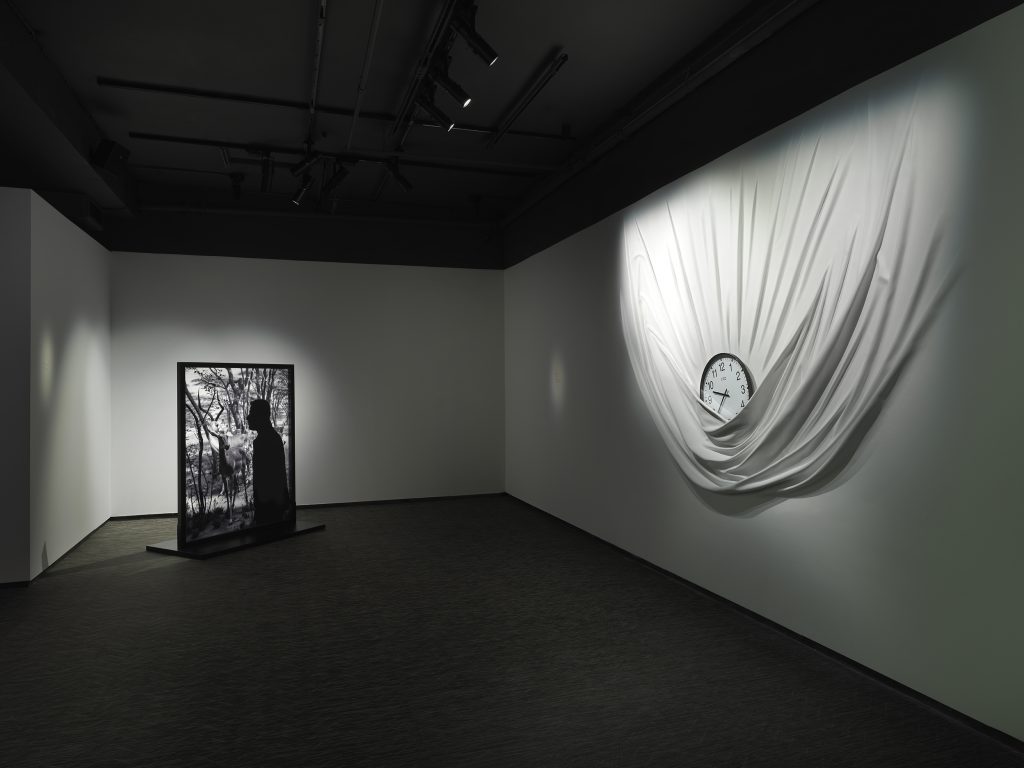 Installation view in a darkened gallery space with an illuminated black and white photograph installed as a standing piece, and a hanging clock in what appears to be white fabric along the wall, by Daniel Arsham.
