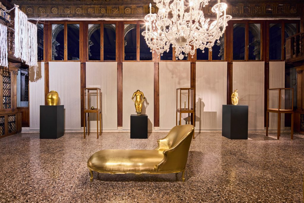 An exhibition in venice inside a historic palazzo, a collonade that has a giant crystal chandelier and a sculpture of a gold chaise lounge and in the background pedestals with golden sculptures of various body parts.