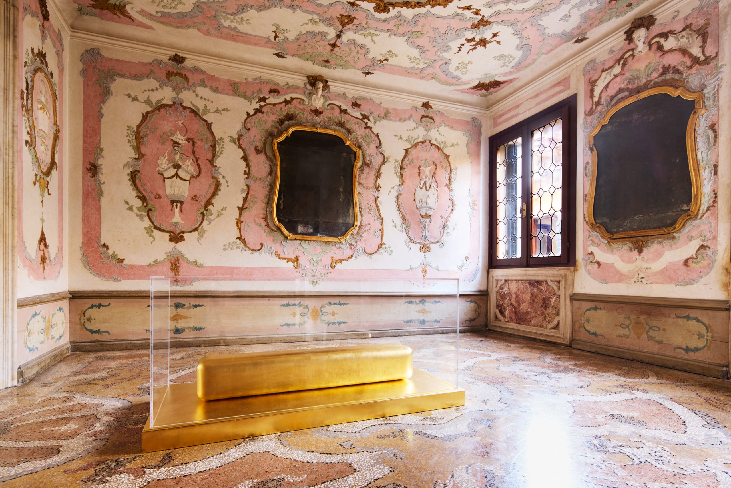 The interior of exhibition in venice in a palazzo that is richly decorated with wall and ceiling paintings in shades of mauve and intricate marble floor. A gold leaf and clear acrylic sculpture rests in the foreground.