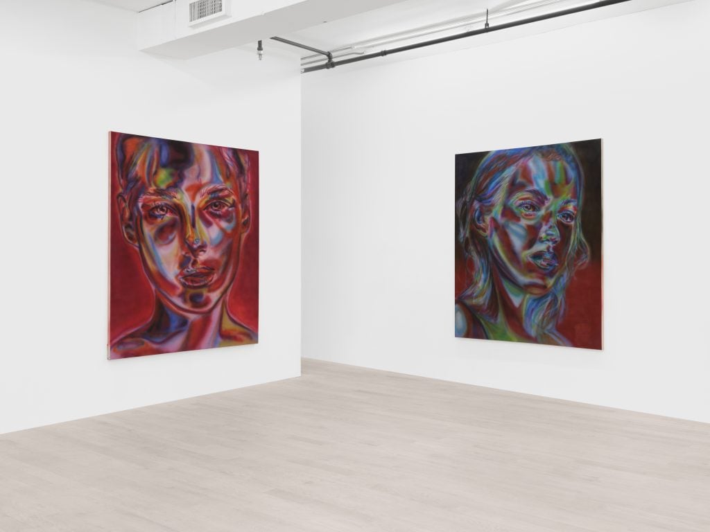Corner of a white gallery space with two paintings by Katie Hector, one on each wall. On the left a headshot portrait in predominantly reds and on the left in predominantly blues. Both have a cyborg aesthetic.
