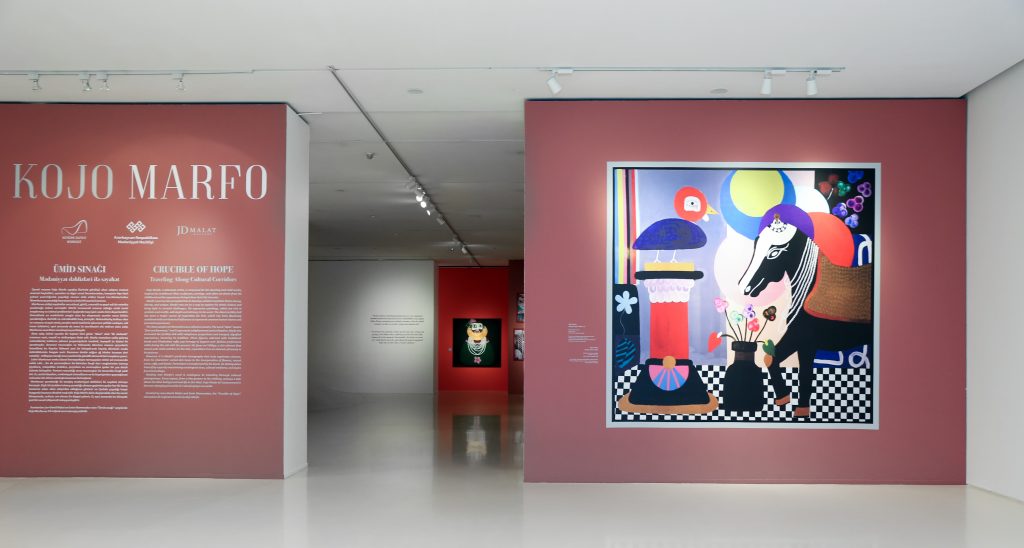 The entrance of the Kojo Marfo retrospective exhibition, with the front wall painted a dusty red, a painting on the right, and didactic vinyl text on the left.