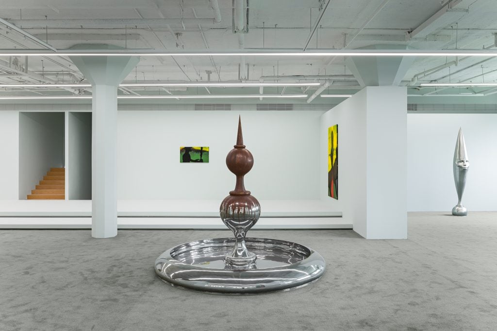 Interior view of a gallery space featuring one symmetrical sculpture in the foreground, two paintings on the back wall, and a silver crescent moon sculpture on the right.