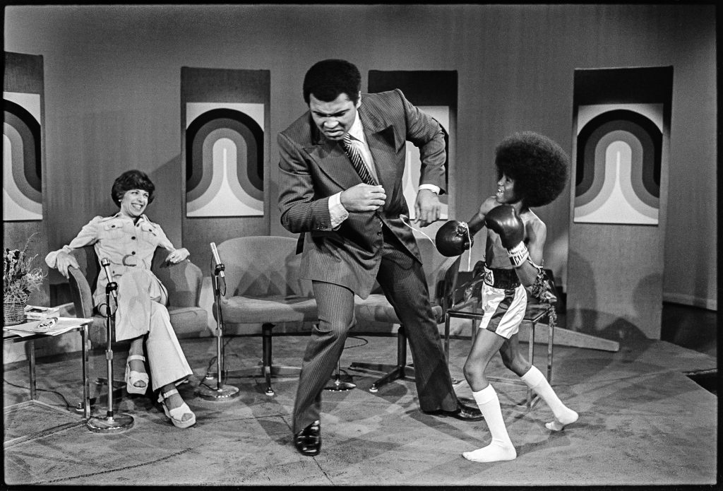 A black-and-white photo of a large man, boxer Muhammed Ali, pretending to be punched by a young child in boxing gloves on a television set