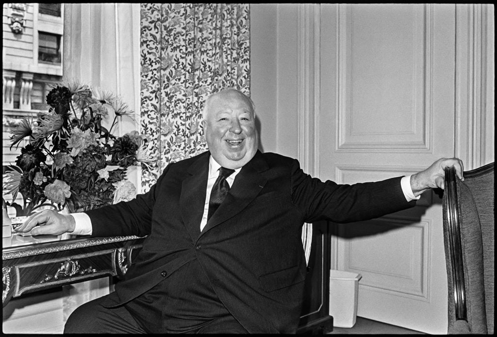 A bald man, director Alfred Hitchcock, smiling while sitting at a desk in a fancy hotel suite