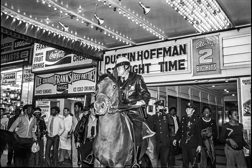 A policeman on a horse outside a 1970s theater, surrounded by other police officers and bystanders