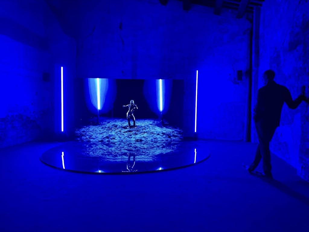 A man looks at a screen in a deep purple gallery, featuring a screen of a figure dancing covered in oil