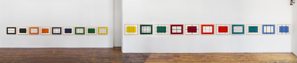 A photograph of twenty colorful geometric prints hung in a long line across two white walls with a deep wood floor below