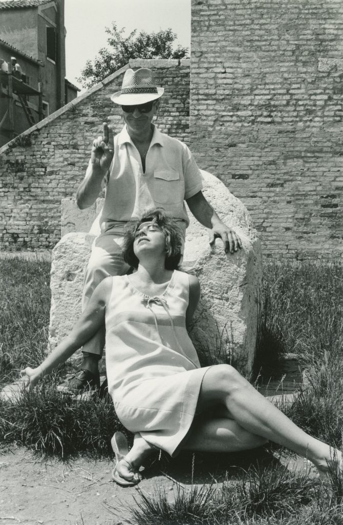 A woman sitting on the ground with her head in the lap of a man who holds up two fingers