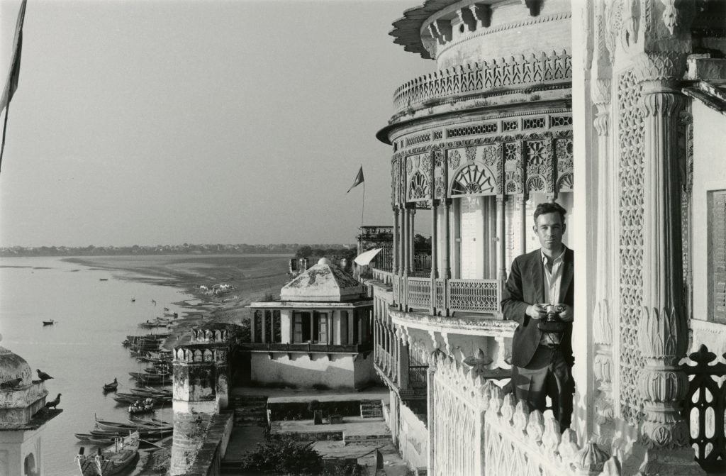 A man standing on a balcony holding a camera, behind him is a view of an Indian city