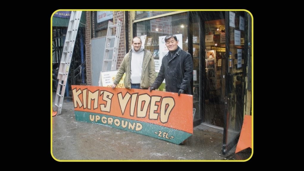 Two man standing outside a store propping up a huge sign, which reads "Kim's Videos."