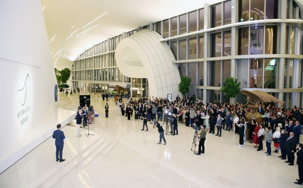 Inside the vaulted white marble, steel, and glass lobby of the Heydar Aliyev Centre, with a crowd of people and cameras watching the opening address of the Kojo Marfo travelling show.