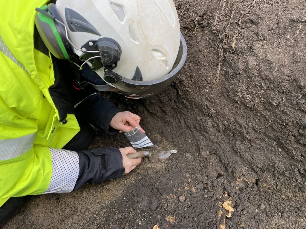 A photograph from above of a woman in protective gear bagging old coins while in the deep dirt hole where they were found