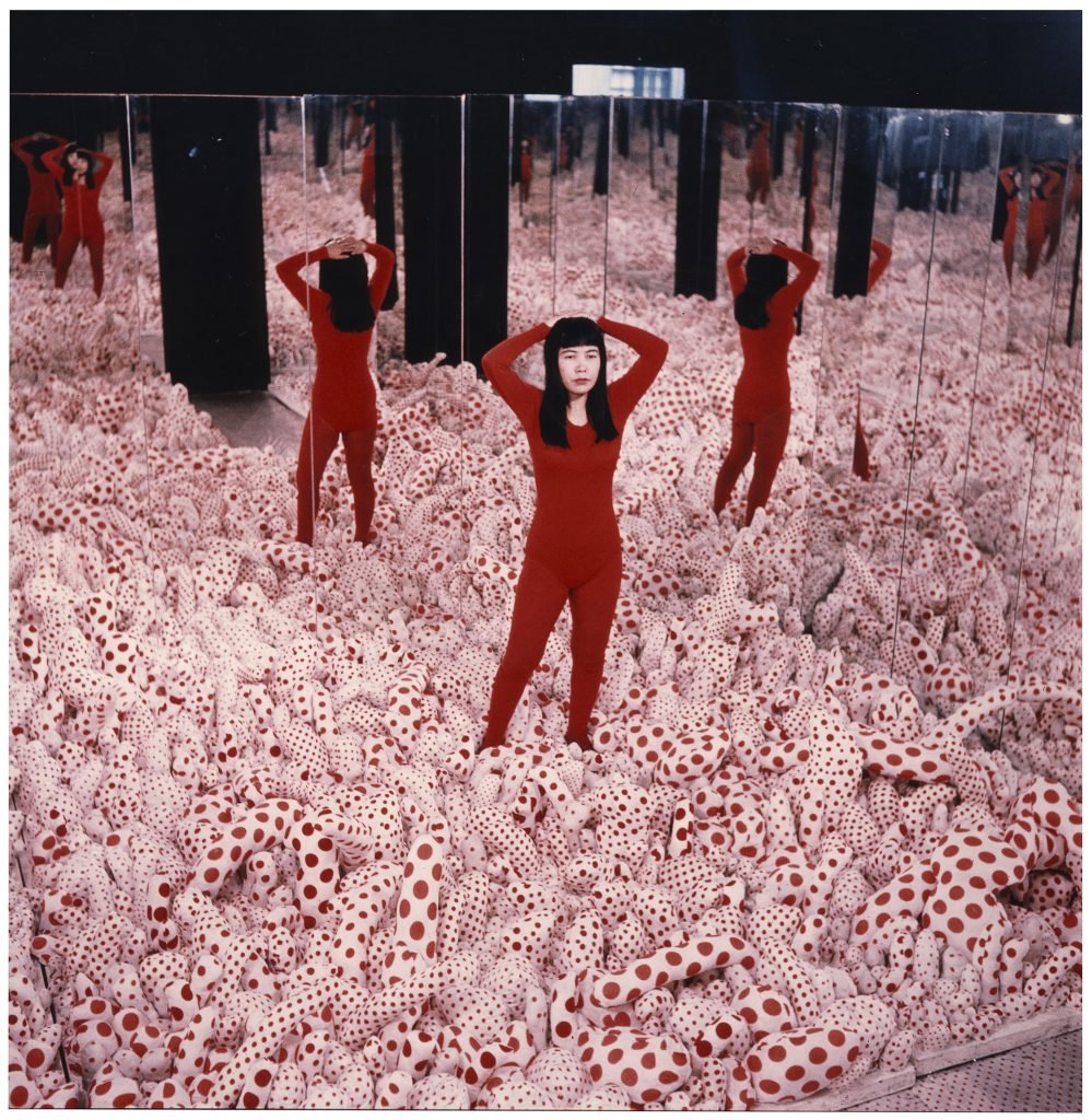A photograph of a 36 year old Yayoi Kusama standing a red jumpsuit at the center of an early infinity room with white abstracted polkadotted forms covering the floor