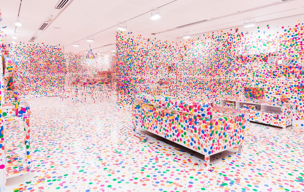 A photograph of a sparsely furnished room painted all white and totally polkadotted in all kinds of bright hues