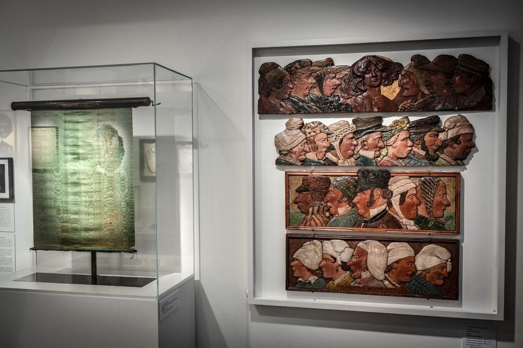 A photograph of two mixed media artworks side by side on a wide wall, including a green-tinged repurposed taxi screen on the right and a wide away of varied faces carved and colored in relief in a case on the right