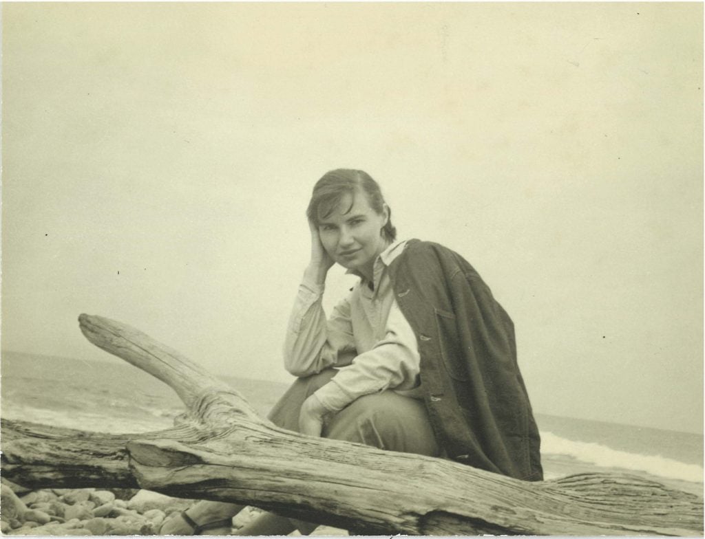 A black and white, sepia-toned photo of a woman posing behind a piece of driftwood, before the ocean