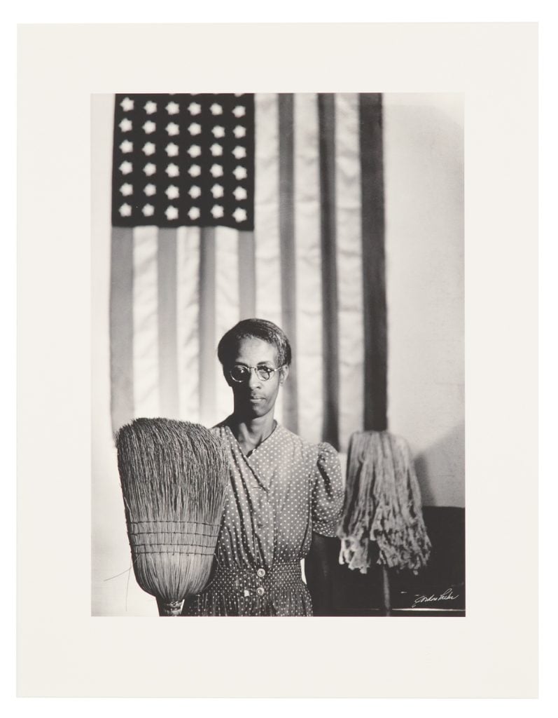 A black-and-white-portrait of a black woman in wire rim glasses and polkadot dress holding a mop and broom standing in front of an American flag in the background.